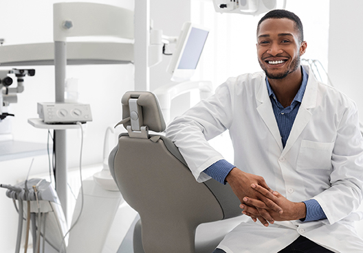 A dentist wearing a white coat and blue shirt sits in the room where he treats patients.