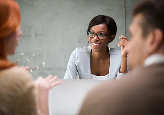 A black female insurance broker wearing glasses and a light grey jacket meets new clients. 