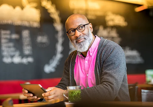 A man with a beard and wearing a pink shirt and gray cardigan holds a tablet in both hands.
