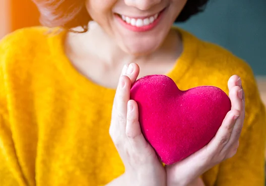 A woman wearing an orange sweater holds a plush red heart in both hands. 