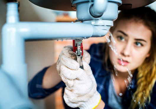 A plumber wearing gloves fixes a leak under a sink at a customer’s home.