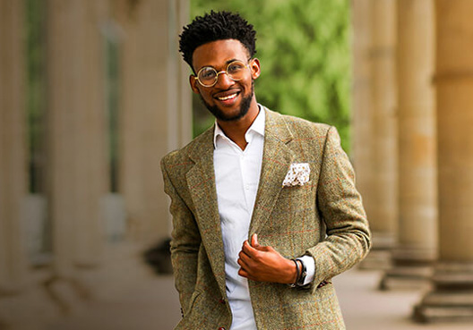 A man wearing a white shirt and brown tweed jacket takes a photo outdoors.