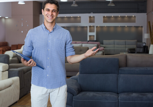 A male business owner wearing a blue shirt welcomes customers to his furniture store. 
