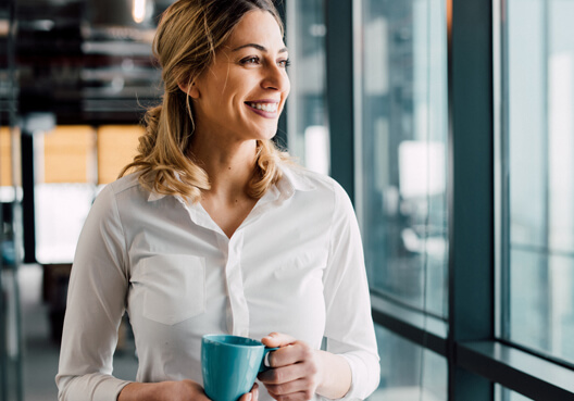 A financial adviser wearing a white blouse holds a blue cup in her hands at her office. A blonde woman wearing a white blouse holds a blue cup in her hands.
