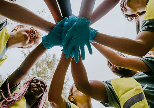 A group of volunteers wearing yellow vests and blue gloves put their hands together.