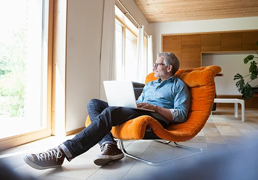 A man wearing jeans and a blue shirt sits in an orange chair with a laptop resting on his lap.