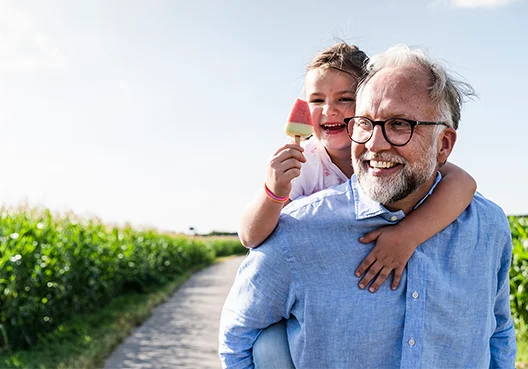 A young girl gets a piggyback ride from her grandfather while she also eats an ice-cream.