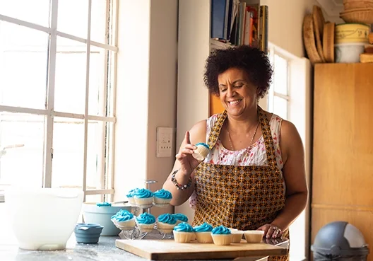 A middle-aged woman wearing an apron bakes cupcakes with blue icing in her kitchen. 