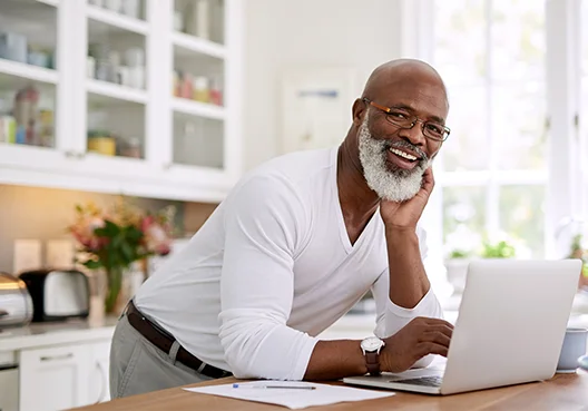 An older man wearing a white shirt uses his laptop in his kitchen. 