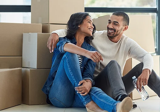 A couple sit on the floor and smile at each other while she holds the keys to their new home.