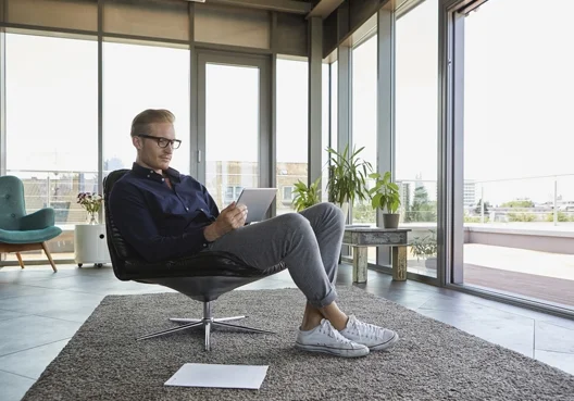 A man wearing glasses and a navy shirt sits in a chair and uses his tablet for online trading. 