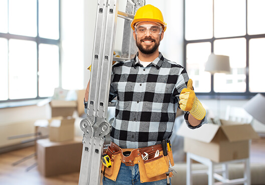 A male contractor dons safety gear and carries a ladder on his shoulder at a worksite. 